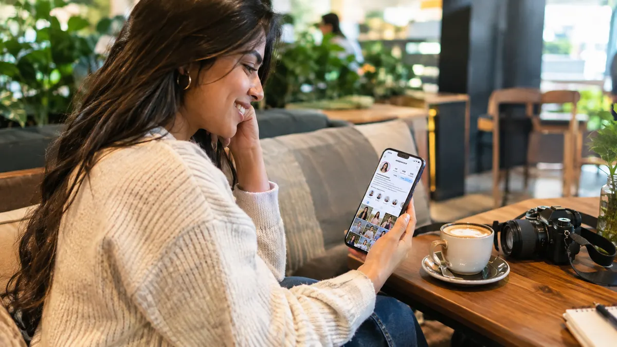 Young woman smiling at her phone showing an Instagram profile with a growing follower count, sitting in a cafe with a camera and coffee beside her