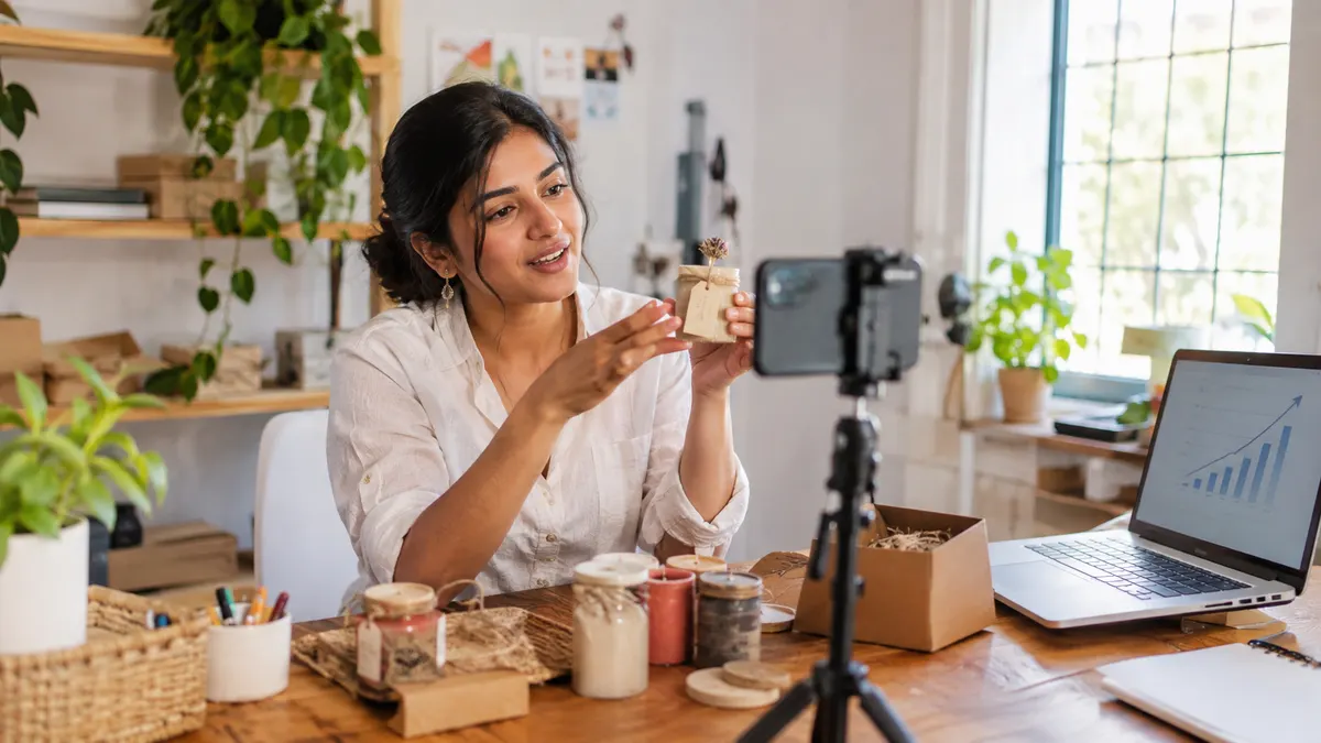 Small business owner filming a product Reel on a phone tripod in a home studio, with a laptop showing a growth chart beside handmade products